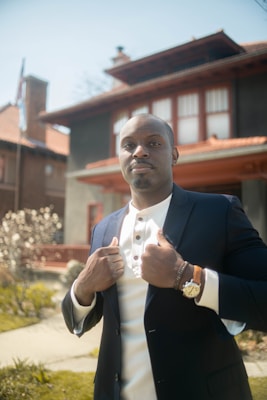 A confident person standing outdoors in front of a large house with a red tiled roof. They are wearing a blue blazer over a white shirt, accessorized with a watch and bracelet. The background includes a well-maintained garden with flowering plants and a path leading up to the house.
