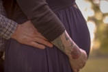 A close-up of a person's hand gently resting on a pregnant belly, with another hand touching the back. One arm is tattooed with colorful designs, including a rose. The clothing is dark, and the background is softly blurred with warm tones.