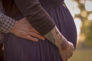 A close-up of a person's hand gently resting on a pregnant belly, with another hand touching the back. One arm is tattooed with colorful designs, including a rose. The clothing is dark, and the background is softly blurred with warm tones.
