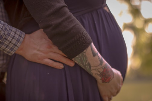 A close-up of a person's hand gently resting on a pregnant belly, with another hand touching the back. One arm is tattooed with colorful designs, including a rose. The clothing is dark, and the background is softly blurred with warm tones.