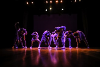 A group of dancers is performing on a stage, each bending forward in a synchronized pose. They are wearing matching outfits, illuminated by dim stage lights creating a dramatic atmosphere. The wooden floor reflects the light, adding depth to the scene.