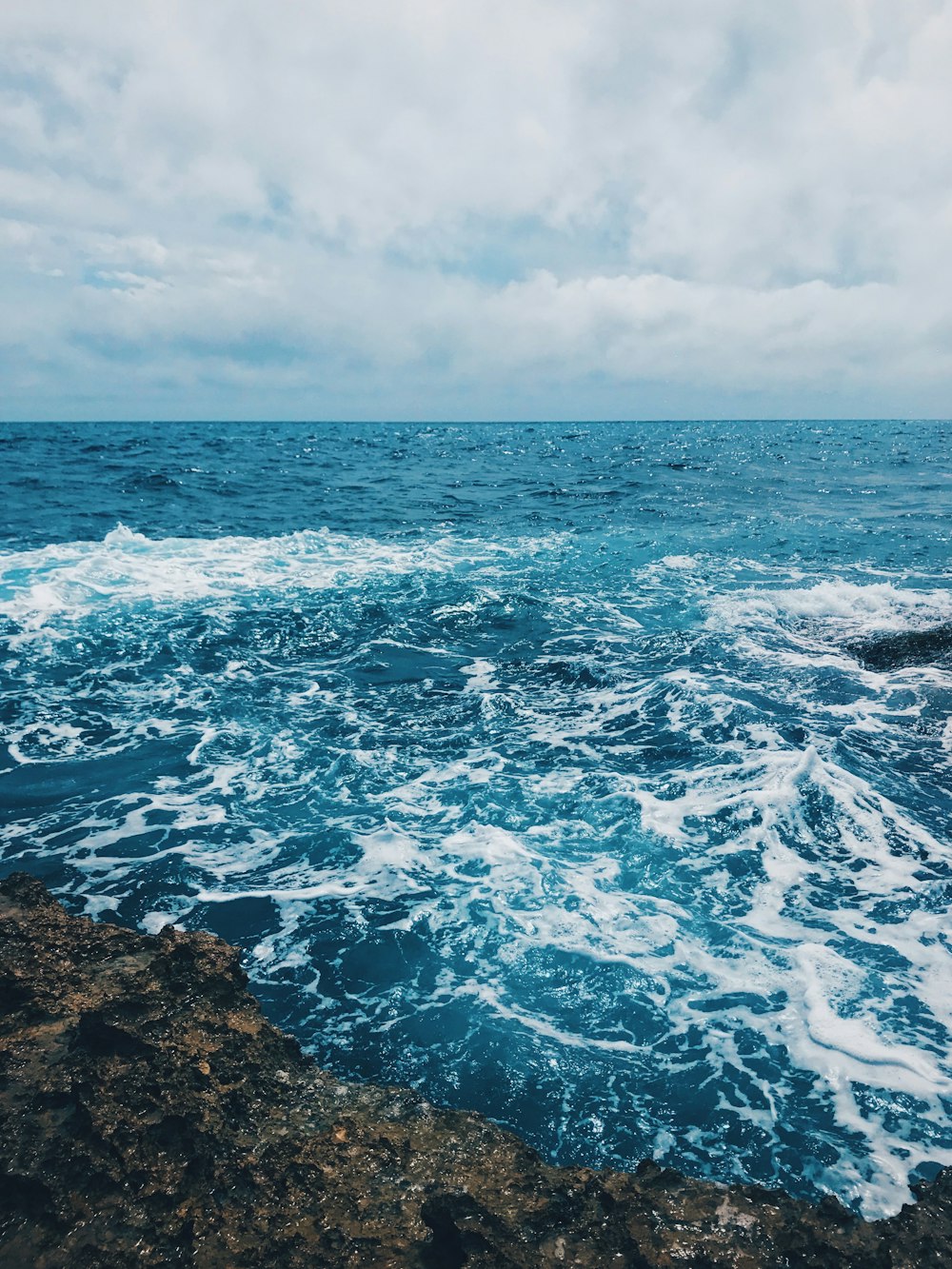 ocean waves crashing on brown rocky shore under white clouds during daytime