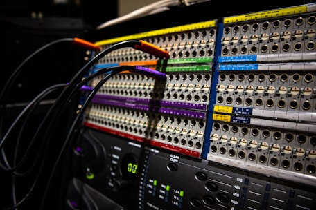 A close-up view of an audio patch panel with multiple rows of connection points. Cables with colorful ends, including orange, purple, and green, are plugged into the panel. Labels and numbers are visible above the connection points.