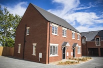 A newly built two-story semi-detached house with a red brick facade and white-framed windows under a clear blue sky. The house features a tiled roof and a front garden area with small shrubs. A paved driveway leads up to the entrance, and another house is partially visible in the background.