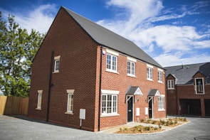 A newly built two-story semi-detached house with a red brick facade and white-framed windows under a clear blue sky. The house features a tiled roof and a front garden area with small shrubs. A paved driveway leads up to the entrance, and another house is partially visible in the background.