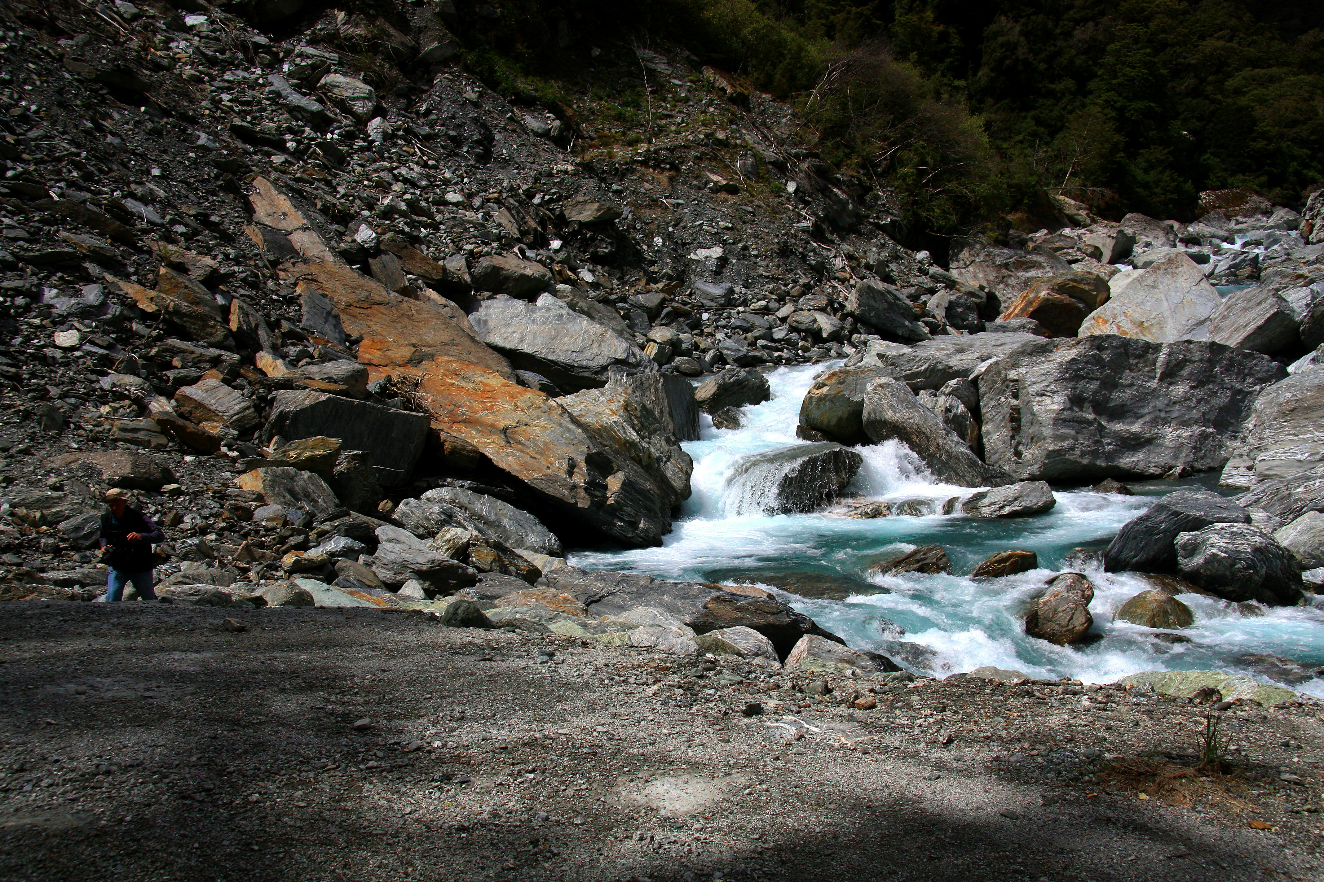 A serene image of children playing near a mountain stream, with a faint border fence in the far background, symbolizing innocent lives near a harsh reality.
