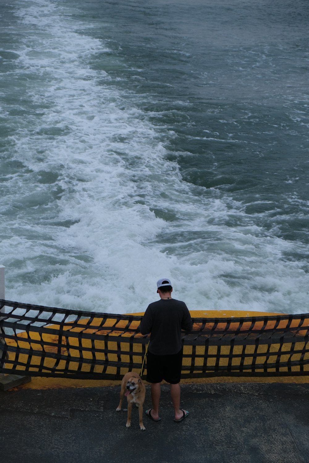 uomo in giacca e pantaloni neri in piedi sul molo di legno marrone vicino alle onde dell'oceano durante il giorno