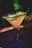 A close-up of a beverage in a martini glass with a salt rim. The drink is yellow with ice cubes and a lime wedge, and there is a stirring stick. The background is dark and slightly out of focus with green and purple lighting adding a vibrant atmosphere.