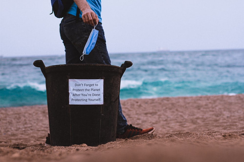 Persona con chaqueta negra sosteniendo un cubo de plástico azul en la playa durante el día