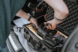 A person is assembling or adjusting a camera inside a protective case in the trunk of a car. The equipment includes a camera body, a monitor, and various cables and accessories. The case has foam padding to protect the contents.