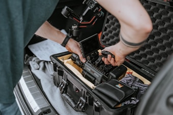 A person is assembling or adjusting a camera inside a protective case in the trunk of a car. The equipment includes a camera body, a monitor, and various cables and accessories. The case has foam padding to protect the contents.