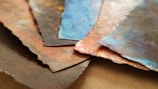 A close-up view of several layered pieces of textured paper with torn edges. The papers display a variety of colors, including earthy browns, deep blues, and hints of red, with a painterly effect.