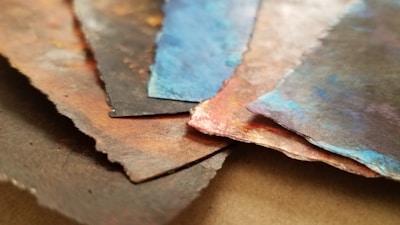 A close-up view of several layered pieces of textured paper with torn edges. The papers display a variety of colors, including earthy browns, deep blues, and hints of red, with a painterly effect.