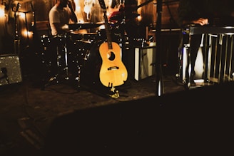 A dimly lit music stage with a focus on a wooden acoustic guitar resting against a drum set. Additional musical equipment, including a keyboard and amplifiers, is visible in the background. Strings of lights create a warm glow across the scene.