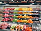 A grocery store display shelf filled with neatly organized fruits, including apples, oranges, grapes, and other assorted fruits. Each type of fruit is arranged in rows with visible price tags. Baskets are placed above the shelf.