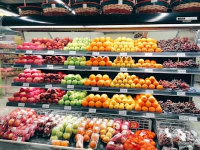 A grocery store display shelf filled with neatly organized fruits, including apples, oranges, grapes, and other assorted fruits. Each type of fruit is arranged in rows with visible price tags. Baskets are placed above the shelf.