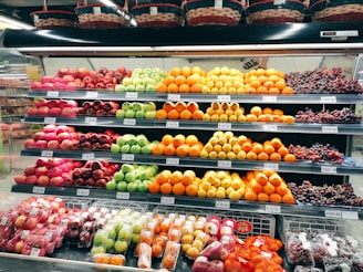 A grocery store display shelf filled with neatly organized fruits, including apples, oranges, grapes, and other assorted fruits. Each type of fruit is arranged in rows with visible price tags. Baskets are placed above the shelf.