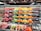 A grocery store display shelf filled with neatly organized fruits, including apples, oranges, grapes, and other assorted fruits. Each type of fruit is arranged in rows with visible price tags. Baskets are placed above the shelf.