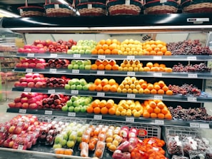 A grocery store display shelf filled with neatly organized fruits, including apples, oranges, grapes, and other assorted fruits. Each type of fruit is arranged in rows with visible price tags. Baskets are placed above the shelf.