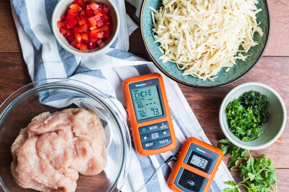A collection of ingredients and tools arranged on a wooden surface include diced red bell peppers in a small bowl, shredded potatoes in a teal bowl, minced herbs in a small green bowl, and raw ground chicken in a glass bowl. Two orange digital thermometers are also present alongside a blue and white striped cloth.
