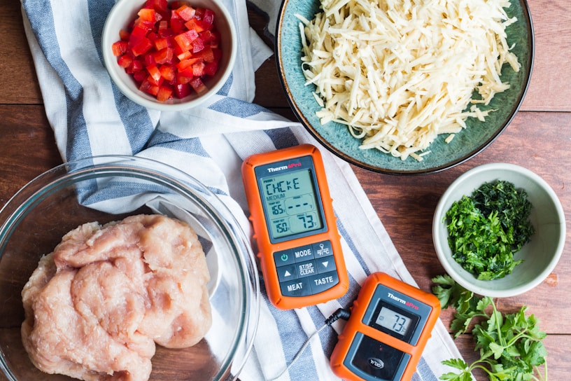 A collection of ingredients and tools arranged on a wooden surface include diced red bell peppers in a small bowl, shredded potatoes in a teal bowl, minced herbs in a small green bowl, and raw ground chicken in a glass bowl. Two orange digital thermometers are also present alongside a blue and white striped cloth.