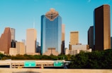 Tall skyscrapers rise prominently against a clear blue sky, displaying a modern architectural skyline. A highway with green traffic signs indicating 'Downtown' and 'I-45 North' runs in the foreground, bordered by lush green trees.