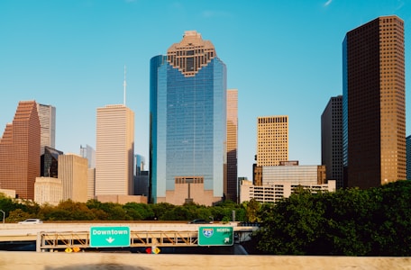 Tall skyscrapers rise prominently against a clear blue sky, displaying a modern architectural skyline. A highway with green traffic signs indicating 'Downtown' and 'I-45 North' runs in the foreground, bordered by lush green trees.