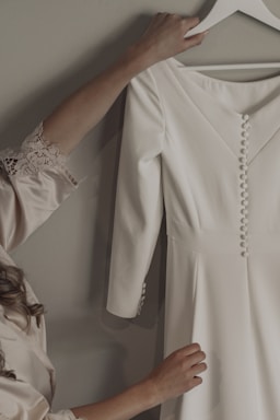A person with long, wavy hair is holding a white dress on a hanger. The dress has long sleeves and a row of buttons down the front. The sleeves also have similar buttons near the cuffs. The person is wearing a garment with lace trim on the sleeve, and appears to be in the process of picking up or adjusting the dress.