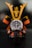 A close-up shot of a wristwatch with a dark face and metallic details, prominently displaying the brand 'Grand Seiko'. The watch rests in front of a decorative figure resembling a samurai with golden and black armor, creating an artistic composition.