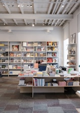 An interior of a modern bookstore or shop featuring shelves filled with various books and magazines. There is a central table displaying more books and items such as stationary or small gifts. The ceiling reveals industrial style with exposed pipes and track lighting. The room is well-lit with natural and artificial light, and the floor is made of multicolored tiles.
