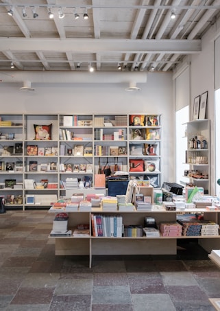 An interior of a modern bookstore or shop featuring shelves filled with various books and magazines. There is a central table displaying more books and items such as stationary or small gifts. The ceiling reveals industrial style with exposed pipes and track lighting. The room is well-lit with natural and artificial light, and the floor is made of multicolored tiles.