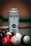 A can of Bira 91 White beer stands upright, with visible condensation droplets on its surface. The can is placed on a surface that appears to be a pool or billiard table, surrounded by an assortment of colorful billiard balls. The background has a warm tone with a soft focus, enhancing the prominence of the can.