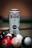 A can of Bira 91 White beer stands upright, with visible condensation droplets on its surface. The can is placed on a surface that appears to be a pool or billiard table, surrounded by an assortment of colorful billiard balls. The background has a warm tone with a soft focus, enhancing the prominence of the can.