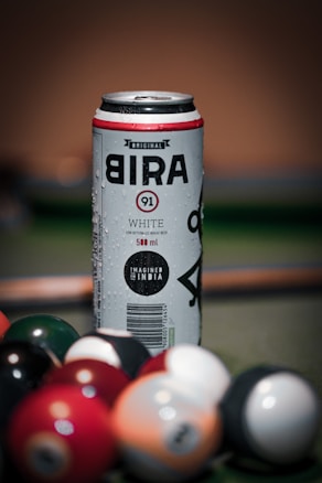 A can of Bira 91 White beer stands upright, with visible condensation droplets on its surface. The can is placed on a surface that appears to be a pool or billiard table, surrounded by an assortment of colorful billiard balls. The background has a warm tone with a soft focus, enhancing the prominence of the can.