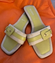 A pair of open-toed sandals with a stylish design, featuring light green leather footbeds and beige straps with a buckle detail. The sandals are set against a bright orange fabric background.