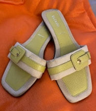 A pair of open-toed sandals with a stylish design, featuring light green leather footbeds and beige straps with a buckle detail. The sandals are set against a bright orange fabric background.