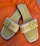 A pair of open-toed sandals with a stylish design, featuring light green leather footbeds and beige straps with a buckle detail. The sandals are set against a bright orange fabric background.
