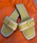 A pair of open-toed sandals with a stylish design, featuring light green leather footbeds and beige straps with a buckle detail. The sandals are set against a bright orange fabric background.