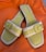 A pair of open-toed sandals with a stylish design, featuring light green leather footbeds and beige straps with a buckle detail. The sandals are set against a bright orange fabric background.