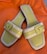 A pair of open-toed sandals with a stylish design, featuring light green leather footbeds and beige straps with a buckle detail. The sandals are set against a bright orange fabric background.
