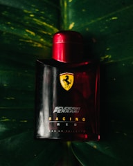 A Ferrari-branded perfume bottle with a bold red cap is positioned against a dark green leaf backdrop. The Ferrari logo and 'Scuderia Ferrari Racing Red' text are prominently displayed on the bottle.