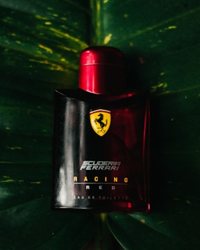 A Ferrari-branded perfume bottle with a bold red cap is positioned against a dark green leaf backdrop. The Ferrari logo and 'Scuderia Ferrari Racing Red' text are prominently displayed on the bottle.