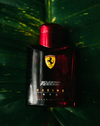 A Ferrari-branded perfume bottle with a bold red cap is positioned against a dark green leaf backdrop. The Ferrari logo and 'Scuderia Ferrari Racing Red' text are prominently displayed on the bottle.