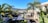 A modern house with large glass windows and a patio surrounded by lush green palm trees and tropical plants. The landscape includes a distant view of mountains under a clear blue sky. The foreground showcases geometric stepping stones set in dark soil, leading to the house.