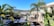 A modern house with large glass windows and a patio surrounded by lush green palm trees and tropical plants. The landscape includes a distant view of mountains under a clear blue sky. The foreground showcases geometric stepping stones set in dark soil, leading to the house.