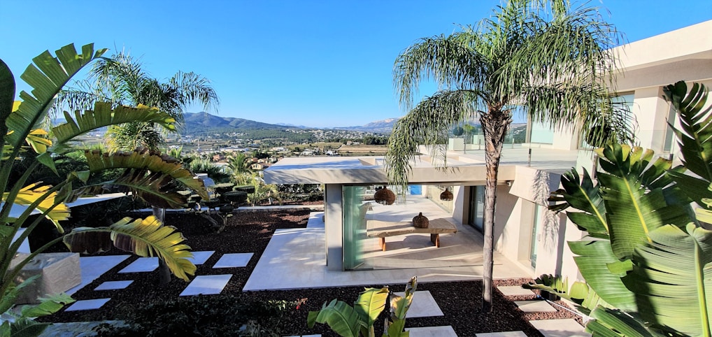 A modern house with large glass windows and a patio surrounded by lush green palm trees and tropical plants. The landscape includes a distant view of mountains under a clear blue sky. The foreground showcases geometric stepping stones set in dark soil, leading to the house.