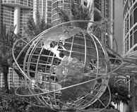 A large metal globe sculpture is positioned in front of modern high-rise buildings with numerous windows. Palm trees and lush greenery surround the structure, creating a contrast between natural elements and urban architecture.