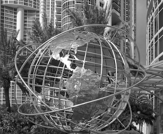 A large metal globe sculpture is positioned in front of modern high-rise buildings with numerous windows. Palm trees and lush greenery surround the structure, creating a contrast between natural elements and urban architecture.
