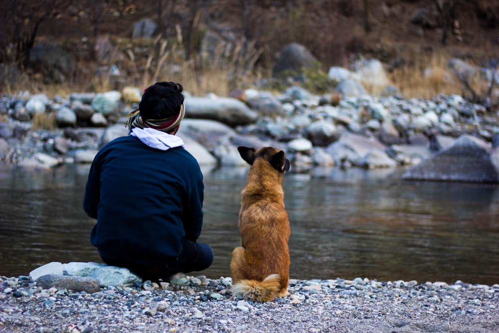 man in black jacket and blue denim jeans sitting beside brown short coated dog on river