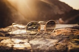 A pair of gold-framed round glasses is placed on a rocky surface, with sunlight creating a warm, golden glow in the background. The glasses are prominently featured and the background appears slightly blurred, providing a serene and calm atmosphere.